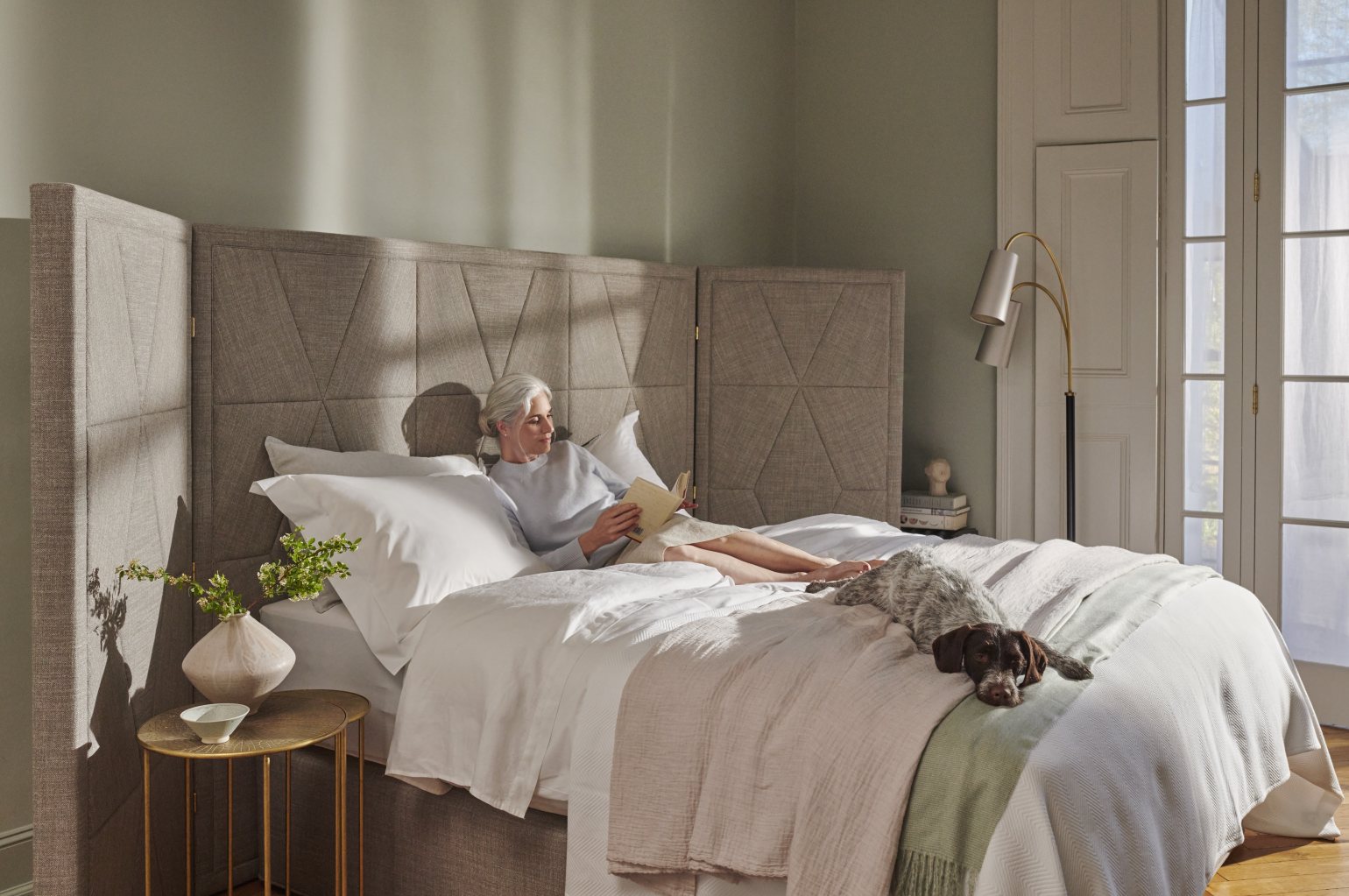 Older woman with her cocker spaniel read and relax quietly in bed with the sun shining through onto the headboard